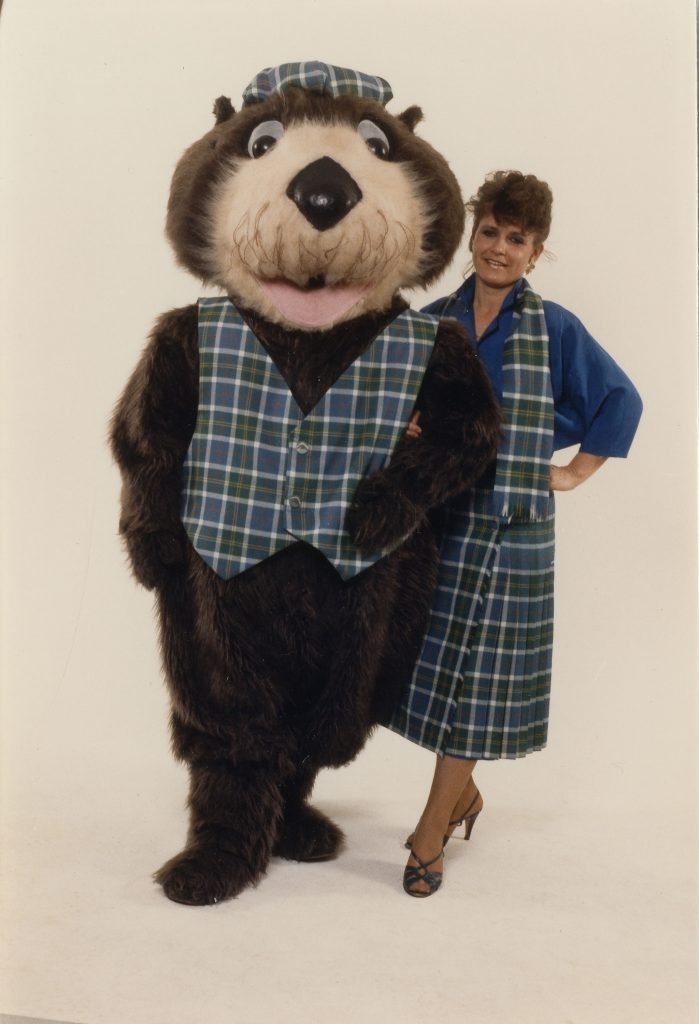 Tillicum the otter mascot and a staff member model Centennial tartan merchandise. Reference code AM1576-S6-12-F39-: 2011-010.2401