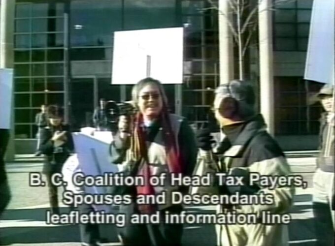 Still from the Saltwater City Television Collective of a Head Tax Payer rally to coincide with the arrival of Prime Minister Paul Martin in Vancouver, November 26, 2005. Sid Chow Tan is holding a megaphone. Reference code: AM1698-S1-: 2023-092.0745